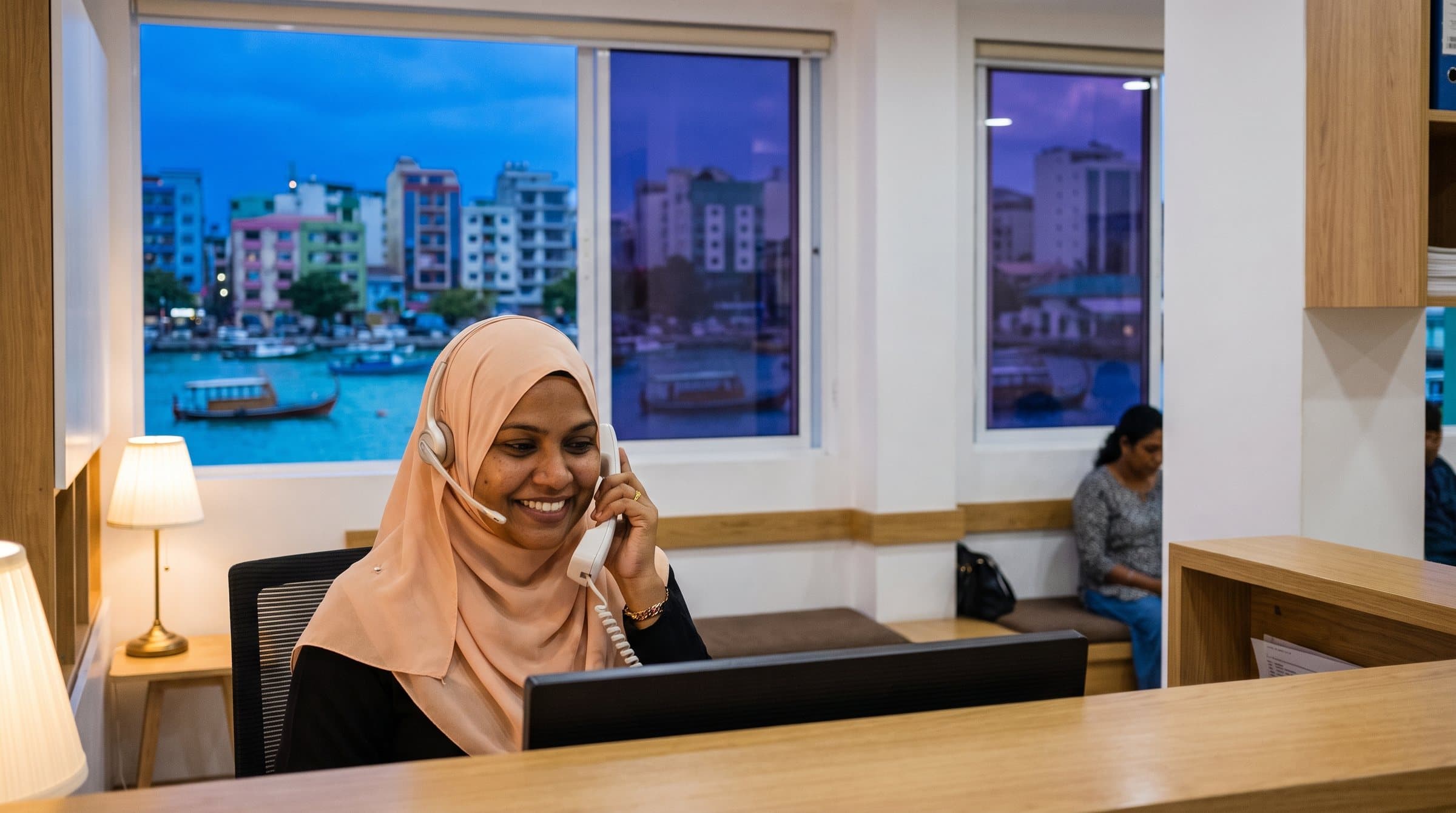 މާލޭގެ ކްލިނިކެއްގެ ރިސެޕްޝަނުގައި ހެޑްސެޓް އަޅައިގެން ފޯނަށް ޖަވާބުދޭ ރިސެޕްޝަނިސްޓެއް، ދޮރުން ފެންނަނީ ހަވީރުގެ ނޫކުލައިގައި ބަނދަރާއި ދޯނިތައް