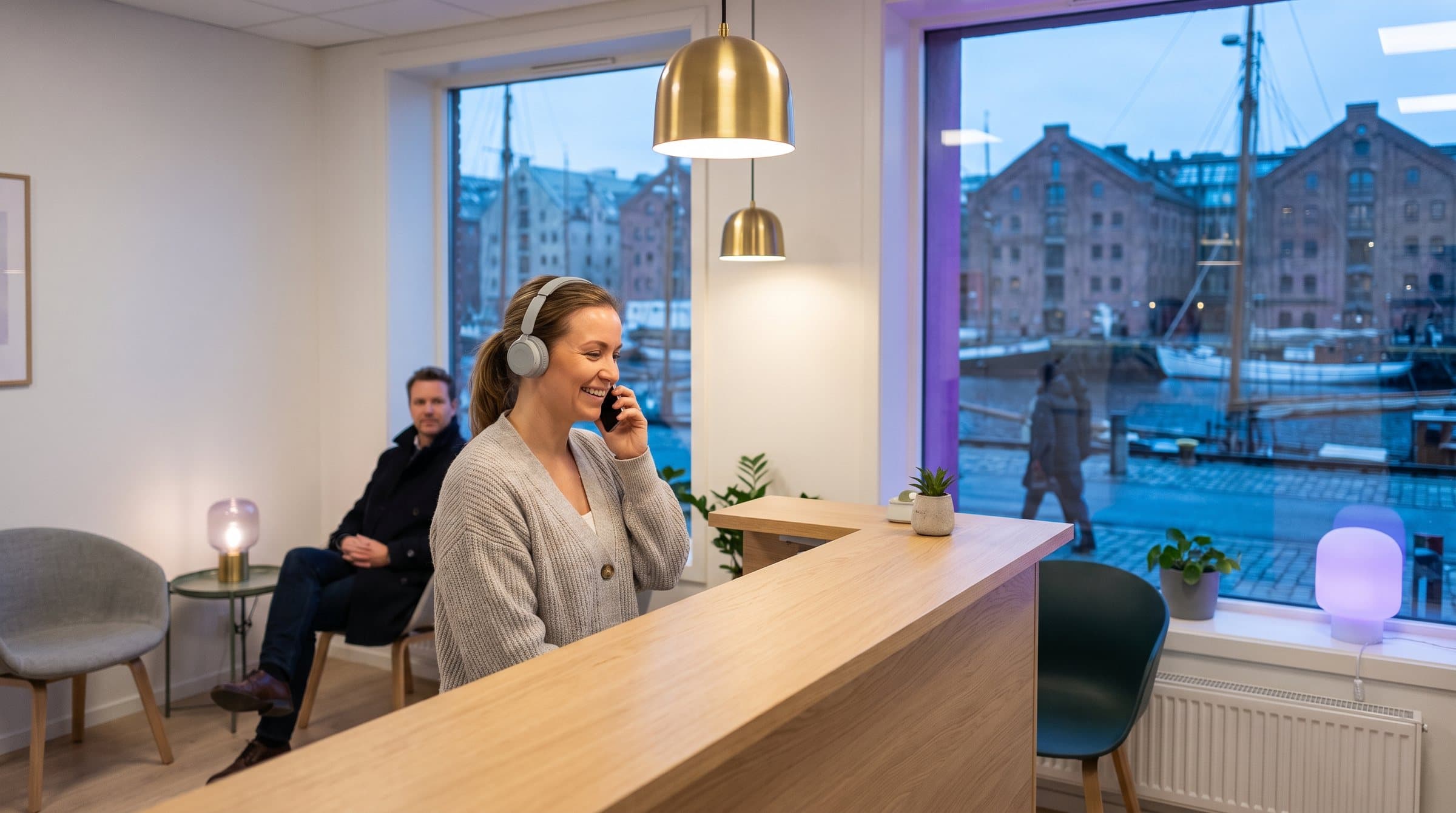 Resepsjonist med headset i telefonen i resepsjonen på en tannklinikk i Oslo, med Oslofjorden og havna i blåtimen utenfor vinduet