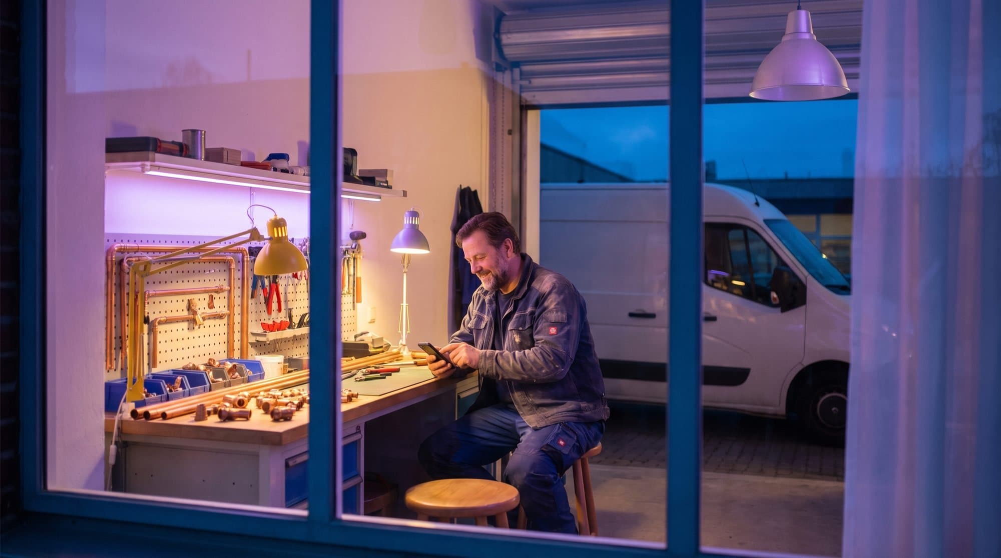 Rørlegger sjekker telefonen i verkstedet sitt ved arbeidsdagens slutt, med varebilen parkert utenfor
